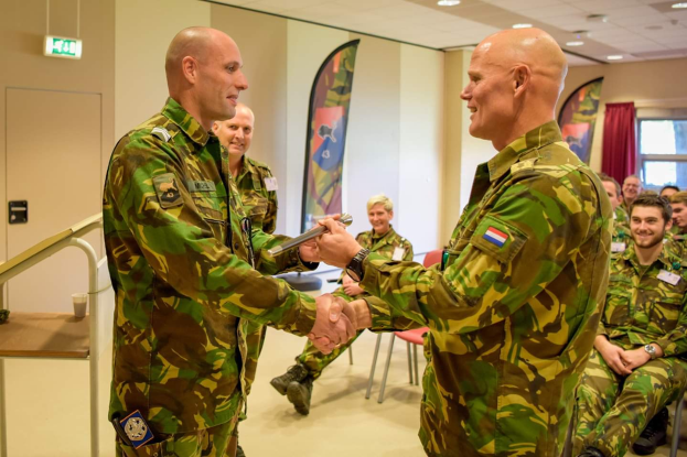 Zwei Männer in Militäruniformen geben sich die Hand vor einer sitzenden Gruppe, mit einem Tisch, Geländer, Fahnen, einer Tür, einem Schild, einem Fenster mit Vorhang und Deckenleuchten im Hintergrund.