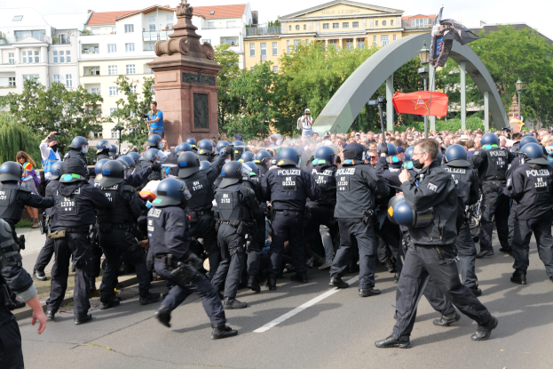 Eine Gruppe von Polizisten in Einsatzkleidung, mit Helmen, geht eine Stra├če entlang, umgeben von einer Menge, mit B├Ąumen, Geb├Ąuden, Laternenmasten, einem Bogen, einer Statue auf einem Sockel und einer Fahne mit einem Mast im Hintergrund unter einem bew├Âlktem Himmel.