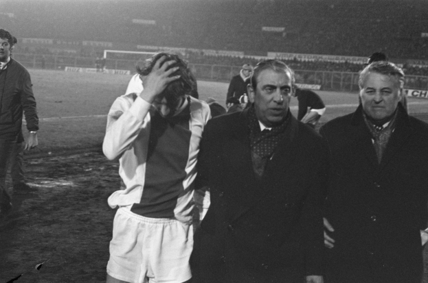 Eine Gruppe von bedrückten Männern in Schwarz-Weiß-Kleidung steht zusammen auf einem Fußballfeld, mit einem Stadion im Hintergrund, wahrscheinlich nach einem gewonnenen Spiel.