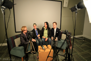 Eine Gruppe von drei Personen sitzt auf einem Sofa und gibt ein Interview. Links sitzt eine Person in schwarzer Jacke und Hose, in der Mitte eine Person in einem Mantel und blauer Krawatte mit einem Mikrofon und rechts eine strahlende Person. In der Mitte steht ein Mikrofonrekorder, Kameras sind auf der rechten und linken Seite und große Scheinwerfer sind vorhanden, im Hintergrund ein Projektionsschirm.