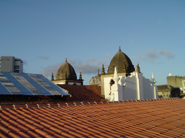 Stadtansicht mit Gebäuden im Vordergrund, Solarpanels auf einem Dach und einem blauen Himmel im Hintergrund.