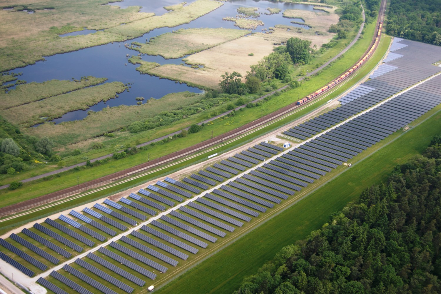 Luftaufnahme einer Solarpark mit Panelen in einem Feld, umgeben von Bäumen, Gras, Pflanzen und Wasser, mit einem Zug auf einer nahen Bahnstrecke.