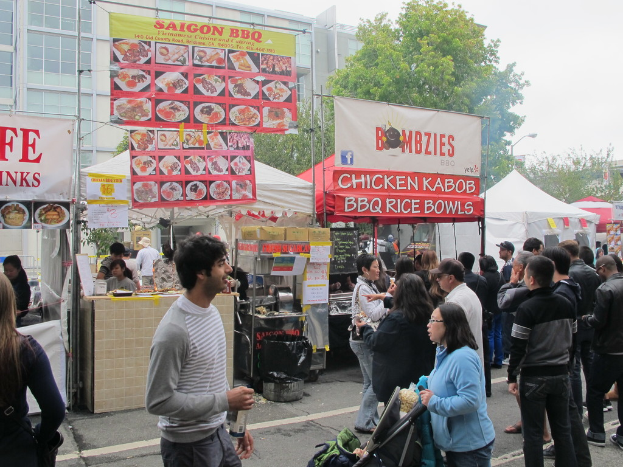 Menschen auf einer Straße vor der Stadt, mit einem Mann in einem grauen und weißen T-Shirt, der eine Wasserflasche hält, und einer Frau in einer blauen Jacke, die einen Kinderwagen schiebt, Stände mit weißen und roten Zelten, bunte Banner und Gebäude, Bäume und eine Straßenlaterne im Hintergrund.