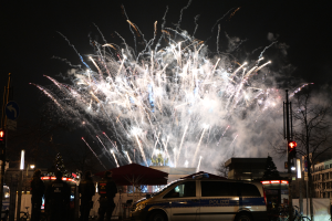 Ein Polizeiwagen vor einer Menschenmenge mit beleuchteten Gebäuden und Bäumen im Hintergrund, unter einem Himmel voller bunter Feuerwerke während einer Silvesterfeier in Berlin.
