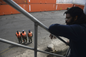 Ein Mann in legerer Kleidung steht auf einem Metallgeländer neben einer Gruppe von Menschen in orangefarbenen Jacken und Mützen, mit Containern und einer Wand im Hintergrund am Hafen von Los Angeles.