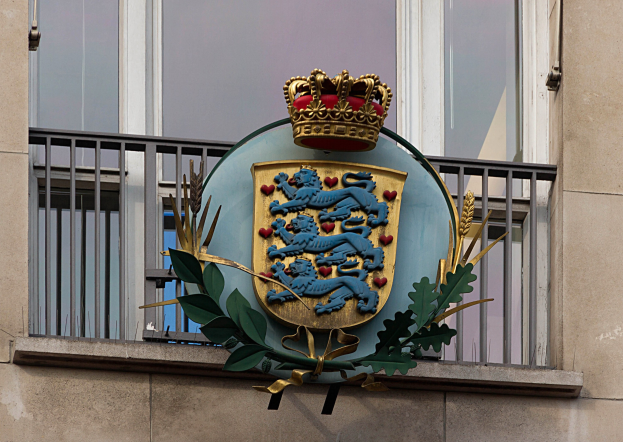 Das Wappen der britischen Königsfamilie auf der Außenseite eines Gebäudes, neben einer Krone und einem Blätter-Logo, mit Fenstern und Gittern im Hintergrund.