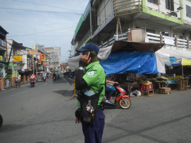 Ein Polizist in einer grün-blauen Uniform und einer blauen Mütze steht auf der Seite einer Straße und spricht auf einem Handy, während hinter ihm Fahrzeuge auf der Straße fahren, Stände mit verschiedenen Gegenständen rechts von ihm und ein bewölkter Himmel mit einem Gebäude mit Fenstern im Hintergrund.