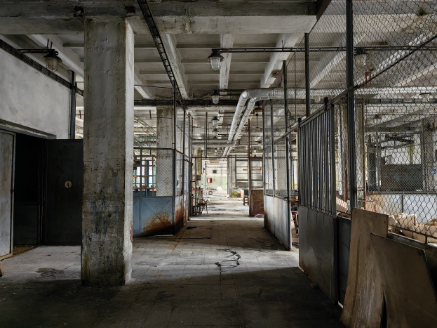 Leerer Raum mit mehreren Fenstern, Säulen, einer Tür, Deckenleuchten, Metallstangen, einem Maschendrahtzaun, verstreuten Gegenständen auf dem Boden und einer Wand, die auf eine verlassene Fabrik hinweist.