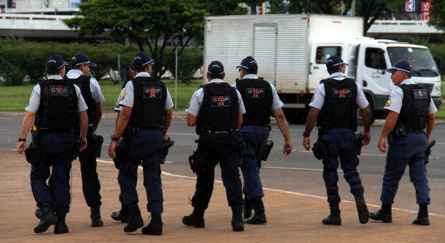 Eine Gruppe von Beamten in Uniform, einschließlich Mützen, Hemden, kugelsicheren Westen, Uhren, Hosen und Schuhen, gehen auf einer Straße. Rechts neben ihnen steht ein Lastwagen in der Nähe eines Zaunes, mit Gebäuden, Bäumen, Pflanzen, Pfählen und Bannern im Hintergrund und einer Brücke oben rechts.