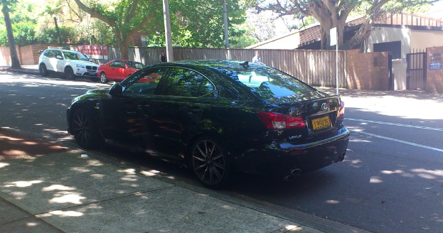 Ein Fußweg führt zu einer Straße mit einem schwarzen Auto und einem weißen und roten Auto, zwei Personen dahinter, Pfosten, einer Umzäunung mit Plakaten und im Hintergrund Bäume, Tore und ein Gebäude mit Wänden und Dächern.