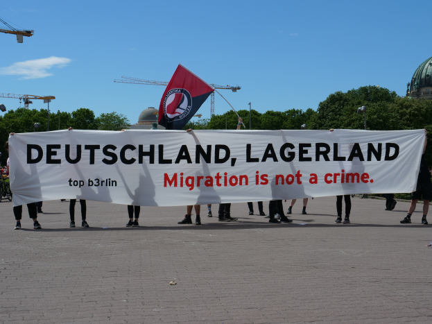 Eine Gruppe von Menschen hält ein Banner mit der Aufschrift "Deutschland, Lagerland Migration ist kein Verbrechen" und eine Fahne, steht auf dem Boden mit Straßenlaternen, Bäumen, Kränen, einem Gebäude und einem bewölkten Himmel im Hintergrund.