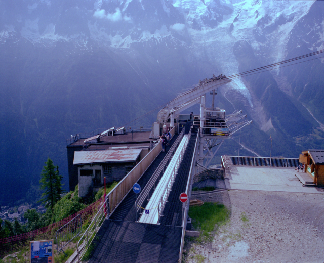 Seilbahn fährt einen schneebedeckten Berg hinauf, mit schneebedeckten Gipfeln im Hintergrund, über eine Straße mit Schildern, Geländern, Grünflächen und Häusern, mit ein paar Menschen sichtbar.