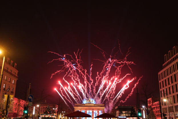 Eine belebte Straßen Szenerie in Berlin an Silvester, gefüllt mit Menschen, Fahrzeugen und Gebäuden, beleuchtet von Feuerwerk und Gebäudelichtern, die eine festliche Atmosphäre schaffen.
