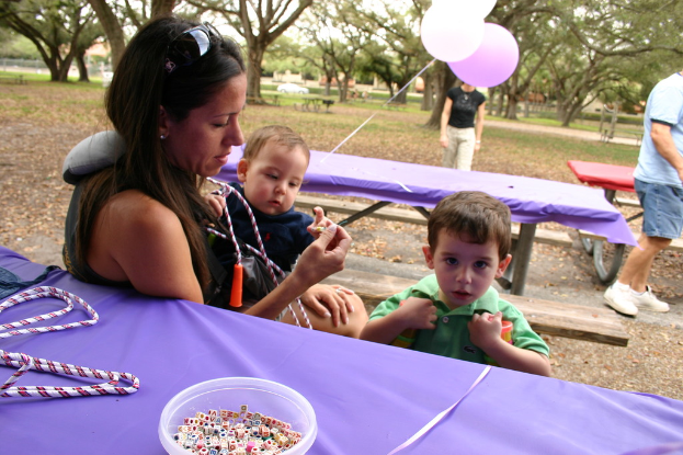 Eine Frau in Schutzbrille hält ein Baby, steht neben einem Jungen in einem grünen Kleid, mit Tischen, einer Violinschrift, einer Schale mit Würfeln und Ballons, während ein Mann rechts steht und Bäume im Hintergrund zu sehen sind.