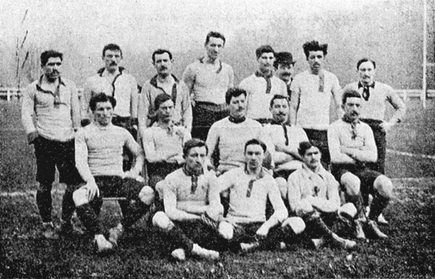 Ein Schwarz-Weiß-Foto einer Fußballmannschaft aus den 1920er Jahren, mit einigen stehenden und einigen sitzend auf dem Boden vor Pfählen, Bäumen und einem klaren Himmel.