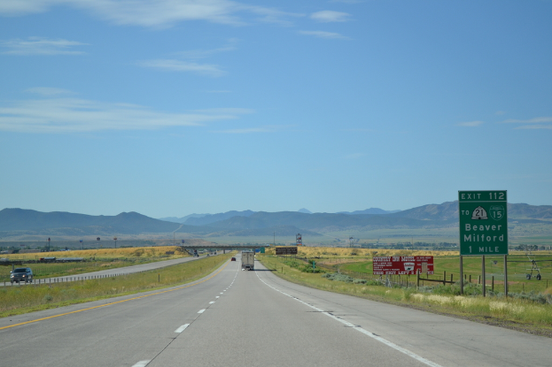 Eine Landschaft mit einem bewölkten Himmel, Hügeln, einer Straße mit Kraftfahrzeugen, Barrieren, Hinweistafeln, Informationsschildern, landwirtschaftlichen Betrieben und Grasflächen.