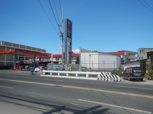 Tankstelle an einer Straßenkreuzung mit parkenden Autos, umgeben von Gebäuden, Strommasten, Schildern, Geländern und Pflanzen unter einem bewölkten Himmel.