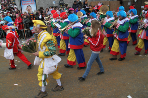 Eine Gruppe bunt kostümierter Menschen marschiert durch eine Straße, spielt Musikinstrumente und hält Blumensträuße, mit Zuschauern und Gebäuden im Hintergrund.