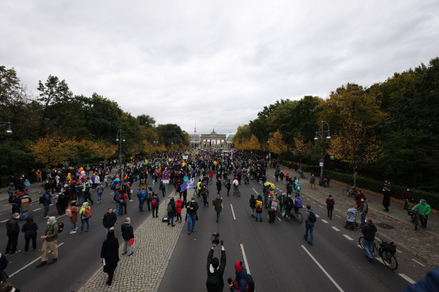 Eine große Gruppe von Menschen geht eine von Bäumen gesäumte Straße in Berlin entlang und hält Kameras während einer Protestdemo, mit einem Gebäude und einem klaren Himmel im Hintergrund.