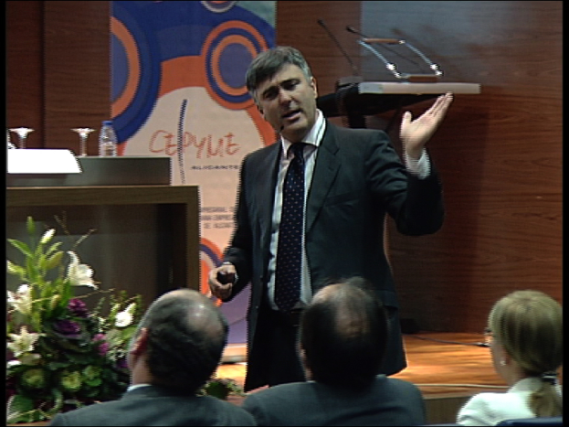 Zwei Männer und eine Frau sitzen auf Stühlen, während ein Mann in einem schwarzen Blazer an einem Podium mit Mikrofonen steht und in ein Handmikrofon spricht, mit einem Blumentopf oder einem Strauß Blumen und einem Tisch mit einem Buch, einer Wasserflasche und anderen Gegenständen in der Nähe, vor einer braunen Wand und einer bunten Tafel mit Text im Hintergrund.