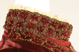 Ein Close-up eines roten und goldenen Kleides auf einer Schaufensterpuppe, das mit feiner Spitzenverzierung gegen einen weißen Hintergrund präsentiert wird.