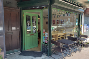 Außensitzbereich des Green Kitchen Vegan Cafe mit Tischen, Stühlen, Pflanzen, einer Fußmatte und einer Glastür mit Plakaten; drinnen sind Tische, Stühle und eine Deckenlampe zu sehen.
