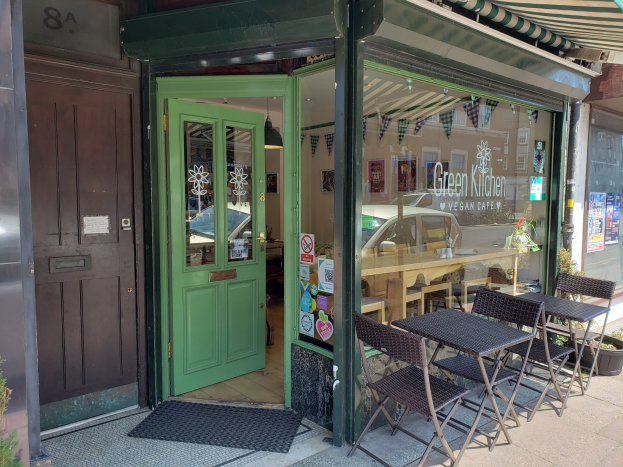 Außensitzbereich des Green Kitchen Vegan Cafe mit Tischen, Stühlen, Pflanzen, einer Fußmatte und einer Glastür mit Plakaten; drinnen sind Tische, Stühle und eine Deckenlampe zu sehen.
