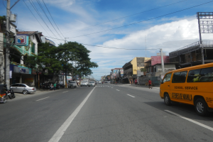 Ein gelber Schulbus fährt eine Straße mit hohen Gebäuden an beiden Seiten entlang, Menschen auf dem Gehweg, Bäume, Strommäste mit Drähten und Schilder mit Text, unter einem bewölkten Himmel.