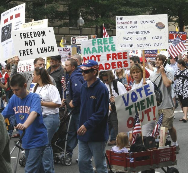 Eine Gruppe von Menschen, die auf einer Straße gehen und Schilder tragen, mit Kindern in einem Wagen vorne und einem Baum, Laternenmast und Gebäude im Hintergrund.