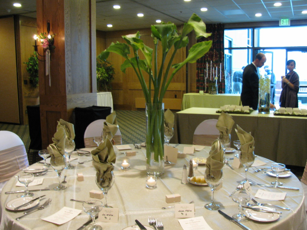 Ein Tisch mit weißer Decke, Tellern, Besteck und einem Glas, umgeben von Stühlen, mit einem grünen Tisch dahinter; ein Mann in einem schwarzen Blazer und eine Frau in der Nähe einer Tür mit zwei Männern dahinter.