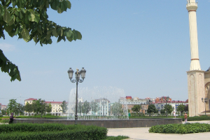 Ein Brunnen in einem Park mit einem hohen Turm im Hintergrund, beleuchtet von Straßenlaternen, mit einer Person im Vordergrund, Bäumen, Gebäuden und einem bewölkten Himmel sowie einem Wasserzeichen auf dem Bild.