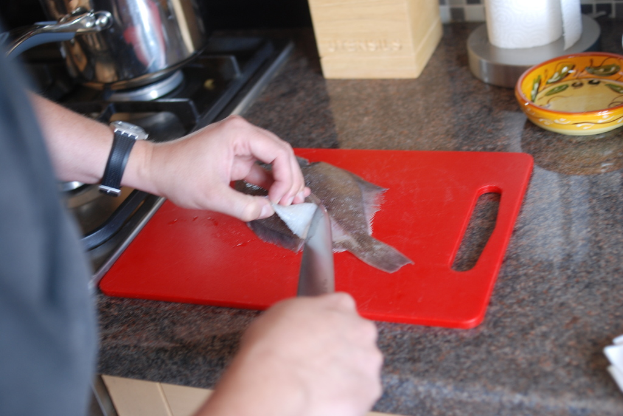 Ein Mann in der Küche, der ein Messer hält und einen Fisch auf einem Schneidbrett auf einer Küchenzeile mit einer Schüssel, Papiertuchrolle, Box, Herd und einem Topf auf dem Herd schneidet.
