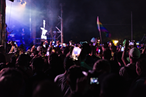 Menge bei einem Konzert mit hochgehaltenen Handys, beleuchtet von Lichtern, umgeben von Pfählen und einer Fahne.
