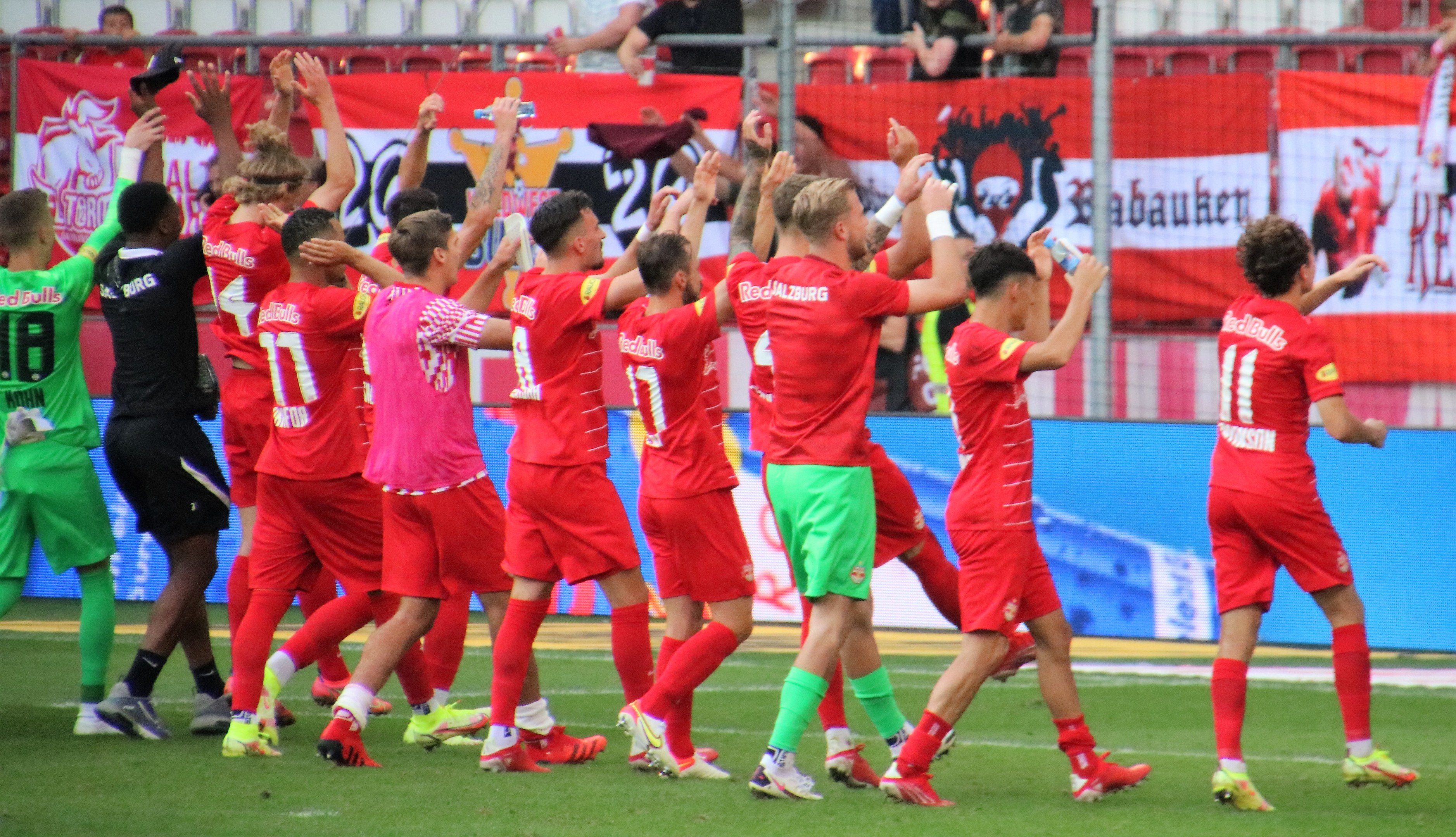 Gruppe von Fußballspielern, die ein Tor feiern, auf einem Feld, einige halten Flaschen und andere klatschen, mit einem Zaun, Bannern, Metallstangen, Stühlen und einer Decke mit Deckenleuchten im Hintergrund.