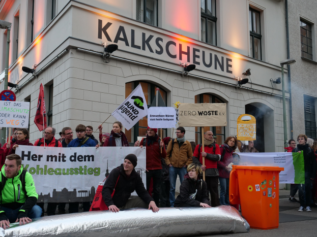 Menschen mit Schildern und Plakaten vor einem Gebäude während einer Demonstration in Deutschland, mit zwei Personen im Vordergrund und einem Müllcontainer auf der rechten Seite.