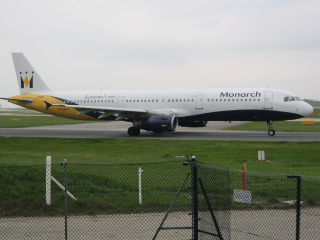 Ein Monarch Airlines Airbus A320-200 auf einer von Gras und einem Zaun begrenzten Start- und Landebahn.