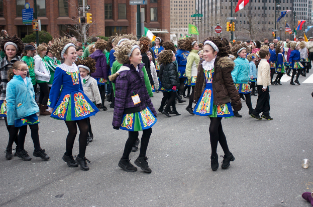 Eine Gruppe von Mädchen in Kleidern mit Jacken und Pullover auf einer Straße tanzend, mit einem roten Gebäude und einem gelben Signalmast dahinter und Gebäuden und Fahnen auf Strommasten im Hintergrund.