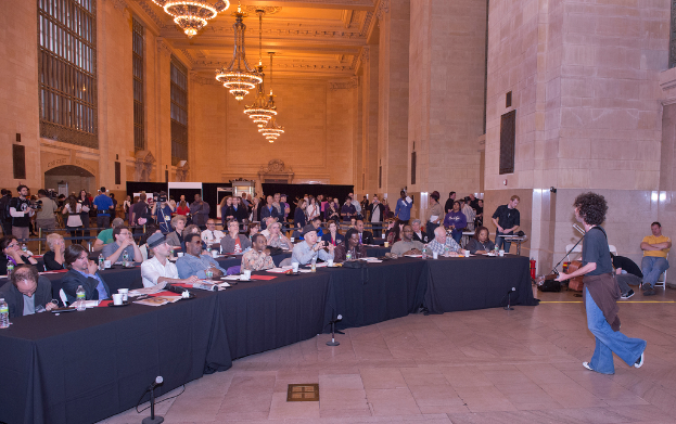 Eine Konzertszene mit einer Menge Menschen, ein Mann spielt Gitarre und singt rechts, eine Wand und Fenster im Hintergrund, Lichter oben und ein Boden unten.