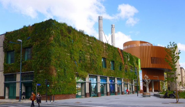 Ein großes Gebäude mit einer grünen Wand an der Seite, umgeben von Straßenlaternen, Verkehrspfosten, Verkehrsampeln, Kraftfahrzeugen, Fußgängern, Bäumen und einem bewölkten Himmel, als Teil des Green Building Project.