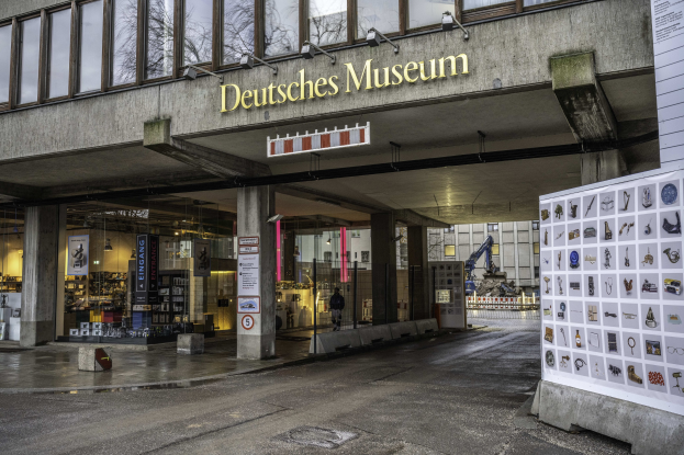 Außensicht des Deutschen Museums in Berlin, ein großer Bau mit Glasfenstern, Säulen und einem Namensschild, neben einer Informationsanzeigetafel, mit anderen Gebäuden, Bäumen und Himmel im Hintergrund.