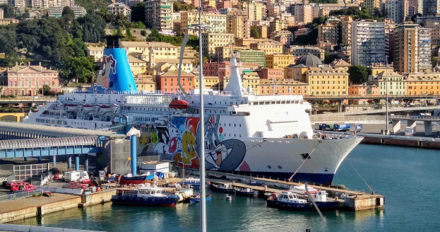 Ein großes Kreuzfahrtschiff liegt in der Nähe einer Stadt, mit einer Brücke auf der linken Seite, Fahrzeugen auf der Straße und Gebäuden und Bäumen im Hintergrund.