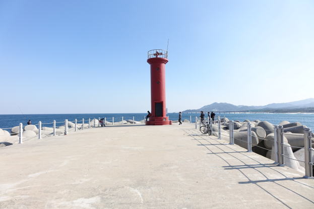 Roter Leuchtturm auf einem Pier am Meer mit Menschen, Fahrrädern, Geländern und Felsen in der Nähe, Hügel und einen klaren blauen Himmel im Hintergrund.