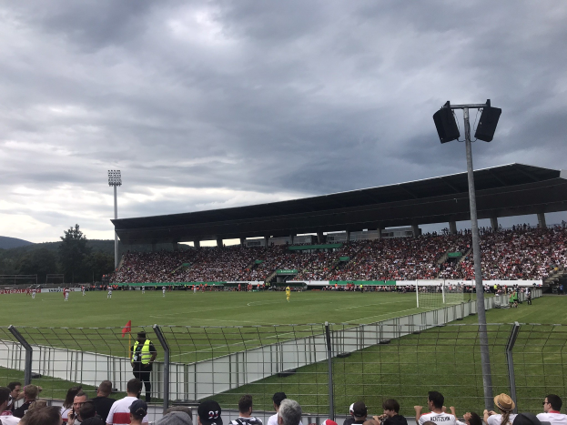 Große Menschenmenge bei einem Fußballspiel in einem Stadion mit bewölktem Himmel und beleuchtetem Feld, umgeben von einem Zaun, Bäumen und Hügeln im Hintergrund.