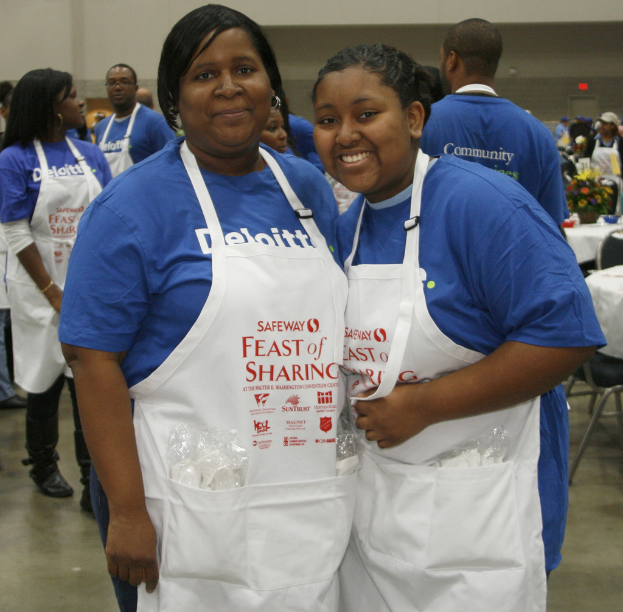 Zwei Frauen in Kochschürzen mit Gegenständen in den Taschen stehen in einem Raum mit Menschen, Tischen, Stühlen, einer Vase und Schüsseln, unter roter Wandbeleuchtung.