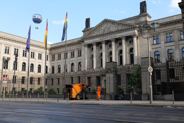 Großes modernes Gebäude mit vielen Fenstern, als Bundestag in Berlin identifiziert, umgeben von Straßeninfrastruktur, Fußgängern und Bäumen unter einem bewölkten Himmel.