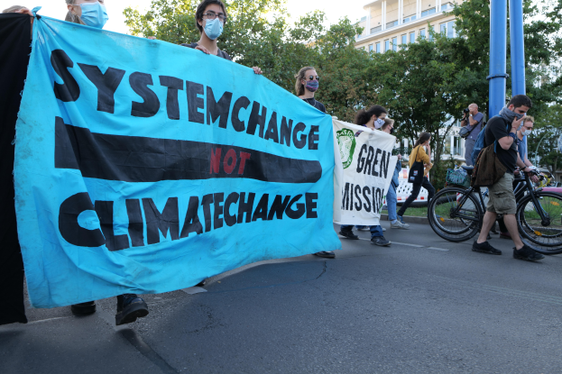 Eine Gruppe von Menschen marschiert mit einem "System Change Not Climate Change"-Schild die Straße entlang, einige tragen Masken und Fahrräder sind in der Nähe geparkt, unter einem klaren blauen Himmel mit Bäumen und Gebäuden im Hintergrund.