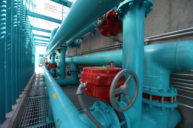 Eine große Gruppe von blauen Rohren und Ventilen in einem Gebäude, mit einem prominenten roten Ventil in der Mitte, Metallstangen links und einer Wand im Hintergrund, die wahrscheinlich den Bau einer Gasleitung darstellt.