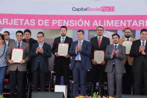 Eine Gruppe von Menschen auf einer Bühne, die klatschen und Zertifikate halten, mit Stühlen in einem Halbkreis darum und einem Banner mit der Aufschrift "Capital Social for Ti" im Hintergrund, zusammen mit Blumensträußen und Rednern unten.