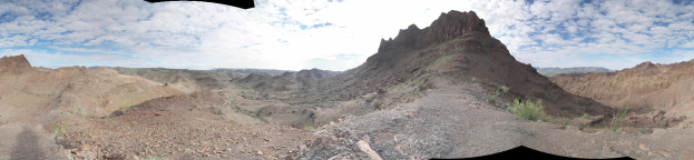 Panoramic view of a mountain range from a hilltop, with hills, vegetation, scattered stones, and a cloudy sky in the background.