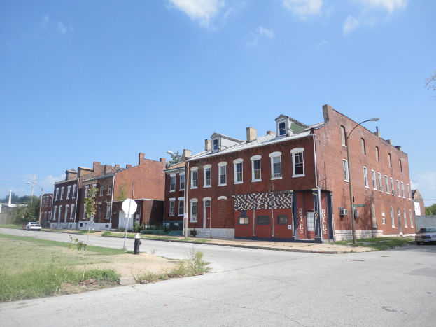Ein Gebäude mit Masten und Straßenlaternen, Bäumen und Fahrzeugen auf beiden Seiten, einem Weg und Gras unten, einem Feuerlöscher und dem Himmel oben.